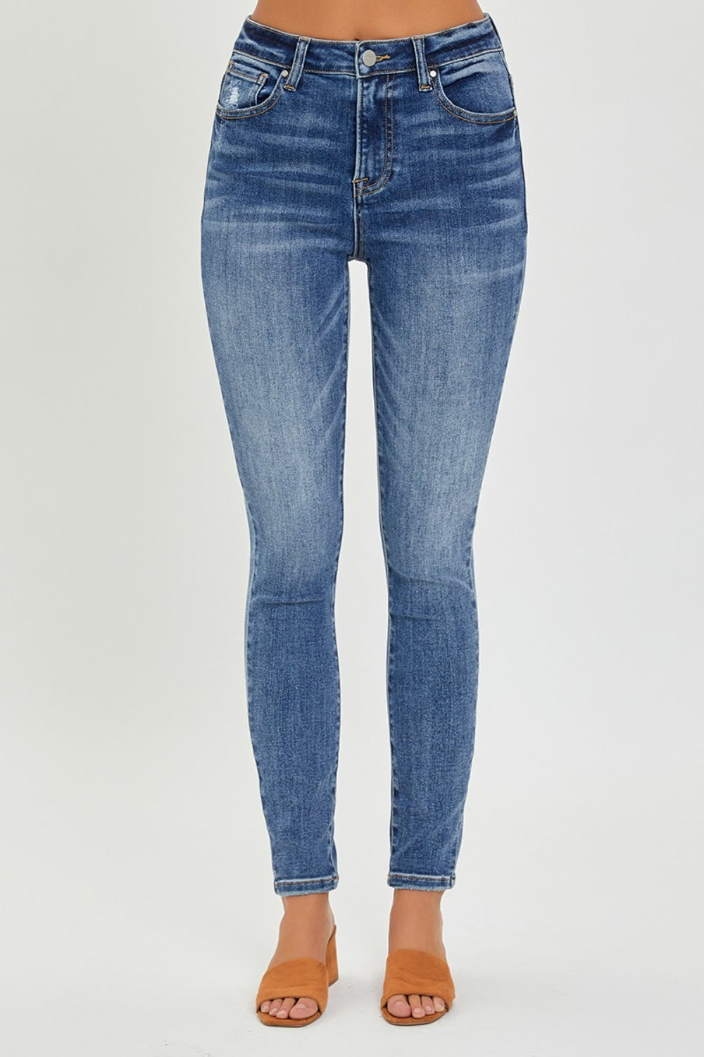 A person models RISEN Full Size Mid Rise Ankle Skinny Jeans standing on a flat surface. The trendy ankle-length design features a button and zip closure, with front and back pockets, paired perfectly with tan open-toe mule shoes.