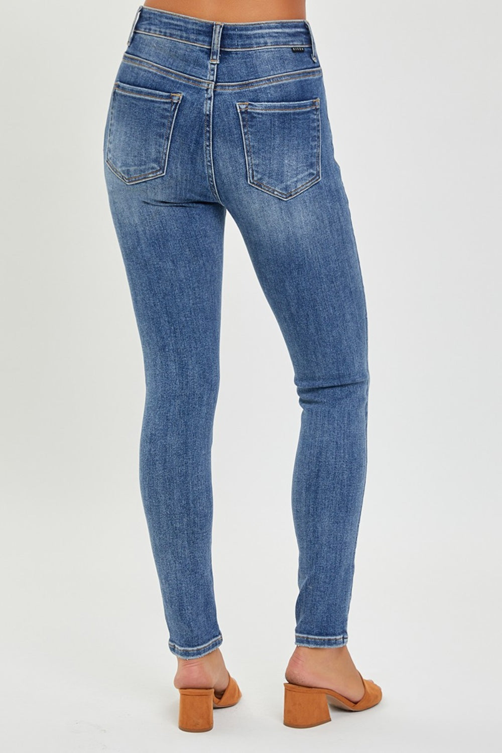 Rear view of a person wearing light blue RISEN Full Size Mid Rise Ankle Skinny Jeans with two back pockets and tan heeled sandals, standing against a plain background. These trendy ankle-length jeans are a versatile wardrobe essential.