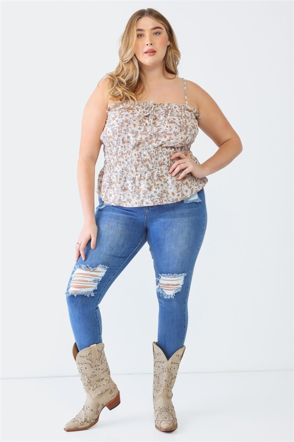 A person in a floral top, distressed blue jeans, and cowboy boots stands against a plain background, wearing a Plus Size Cami | Zenobia Frill Floral Square Neck beneath the vibrant blouse.