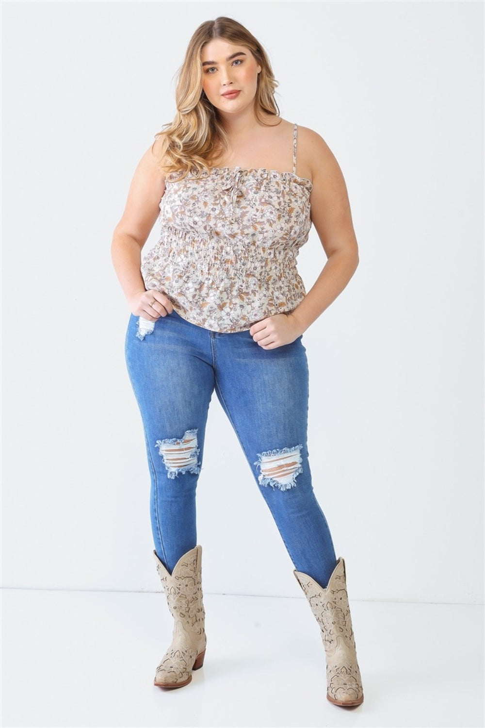 A person stands against a plain backdrop wearing a Plus Size Cami | Zenobia Frill Floral Square Neck under a floral top, paired with ripped blue jeans and cowboy boots, with hands resting in the pockets of their jeans.