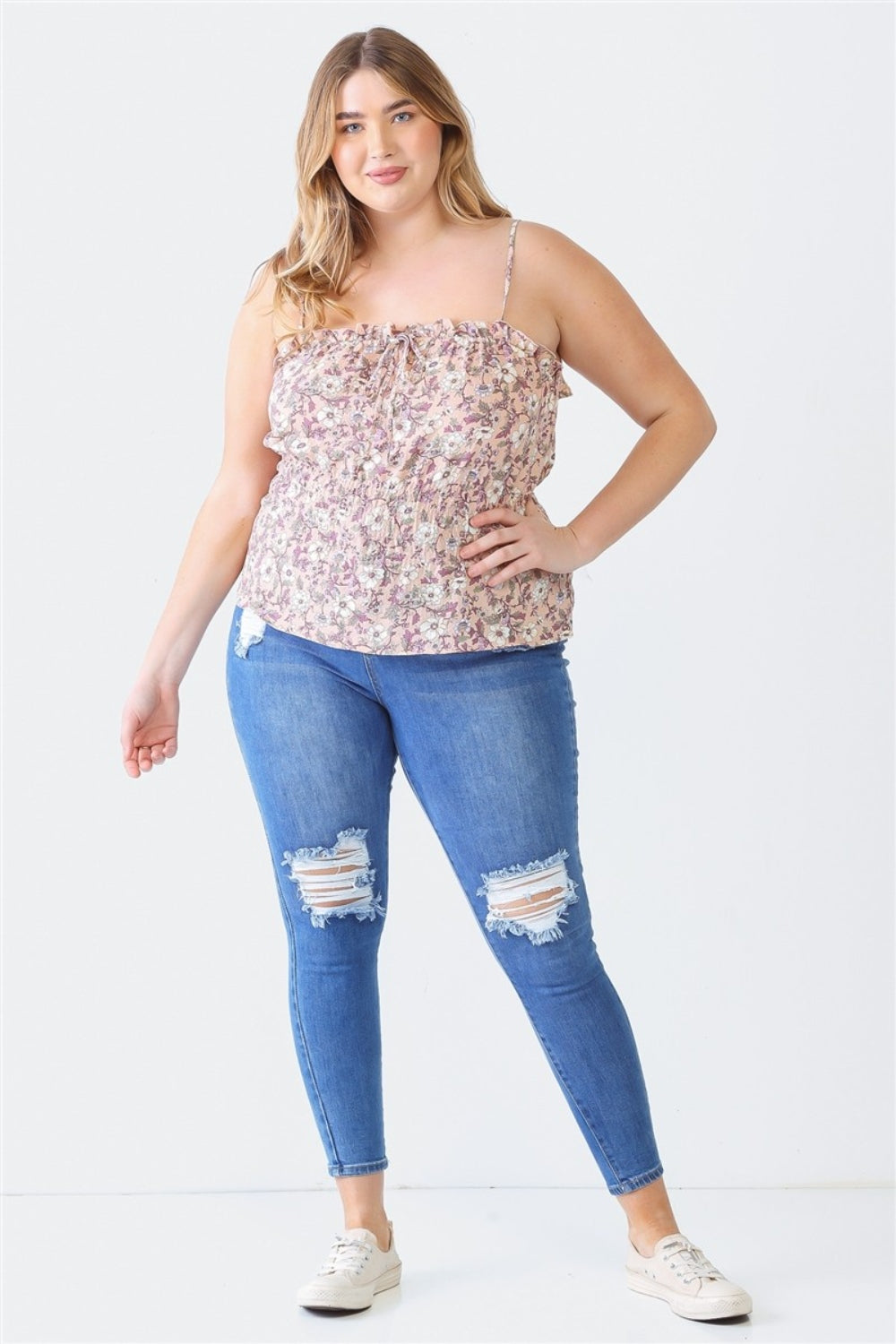 A woman wearing a floral sleeveless top and ripped blue jeans stands against a plain white background. She has long, wavy blonde hair and is wearing white sneakers. Her Plus Size Cami | Zenobia Frill Floral Square Neck perfectly complements her casual, chic look.