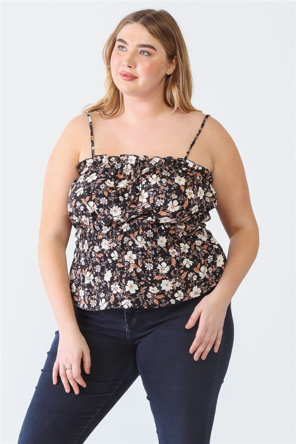 A woman wearing a floral-patterned sleeveless top and dark jeans stands looking off to the side against a plain white background, showcasing her Plus Size Cami | Zenobia Frill Floral Square Neck with confidence.